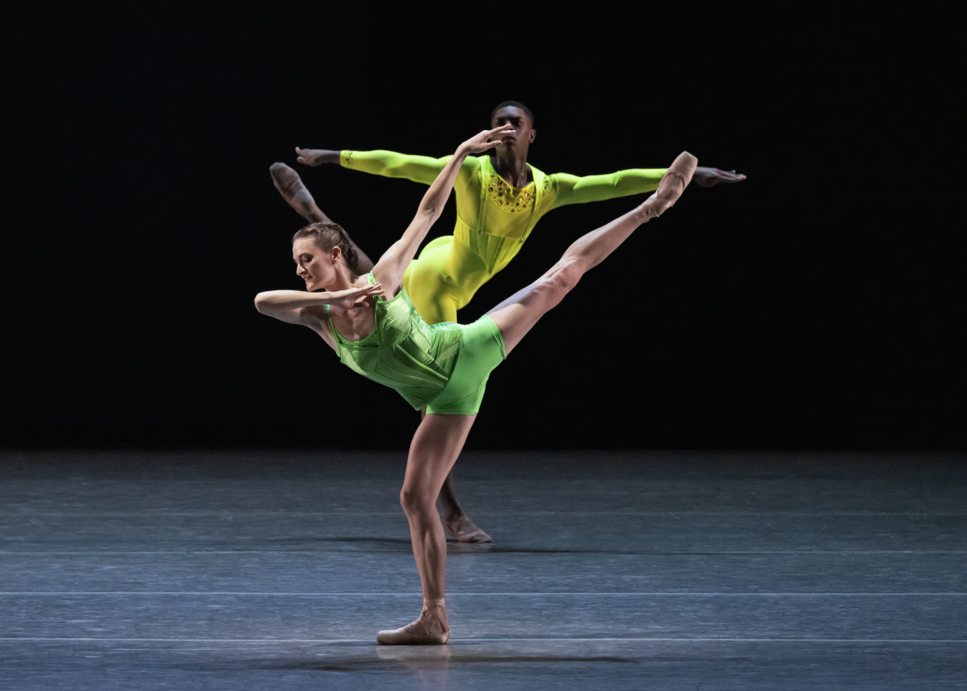 A female dancer in green shorts and a bodice extends her leg in a high arabesque in profile as one hand grazes the shoulder of her opposite arm. Behind her, a man in a yellow long sleeve shirt extends his arms in a T.